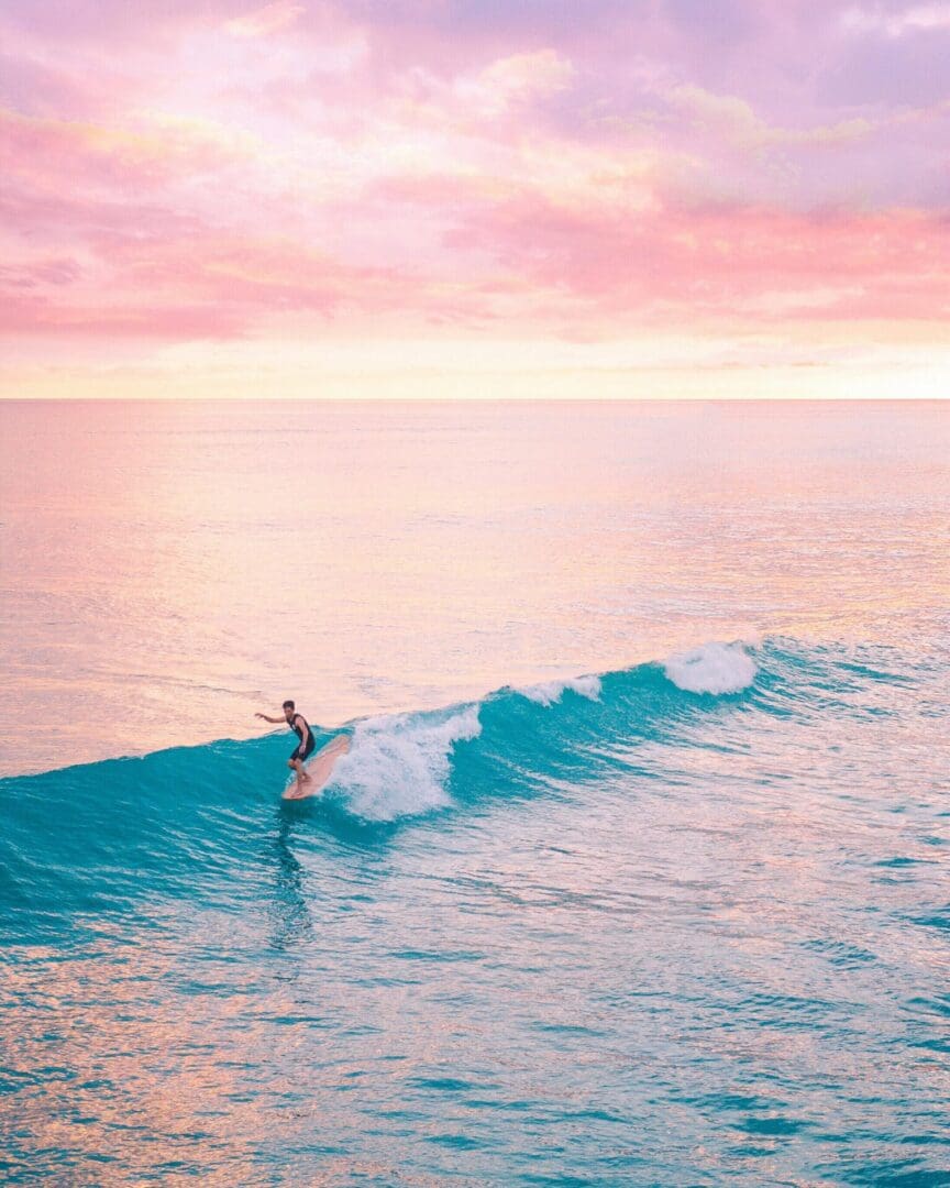 Surfer riding a wave at sunset with a pink sky.